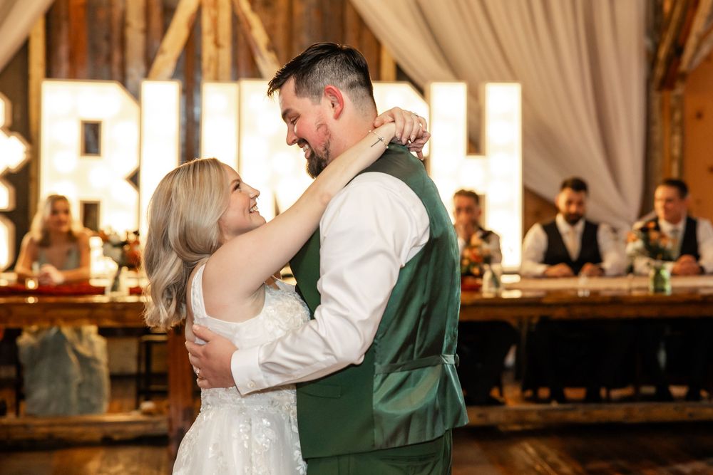 Bride and groom share their first dance in a rustic barn wedding venue with glowing marquee letters in the background.