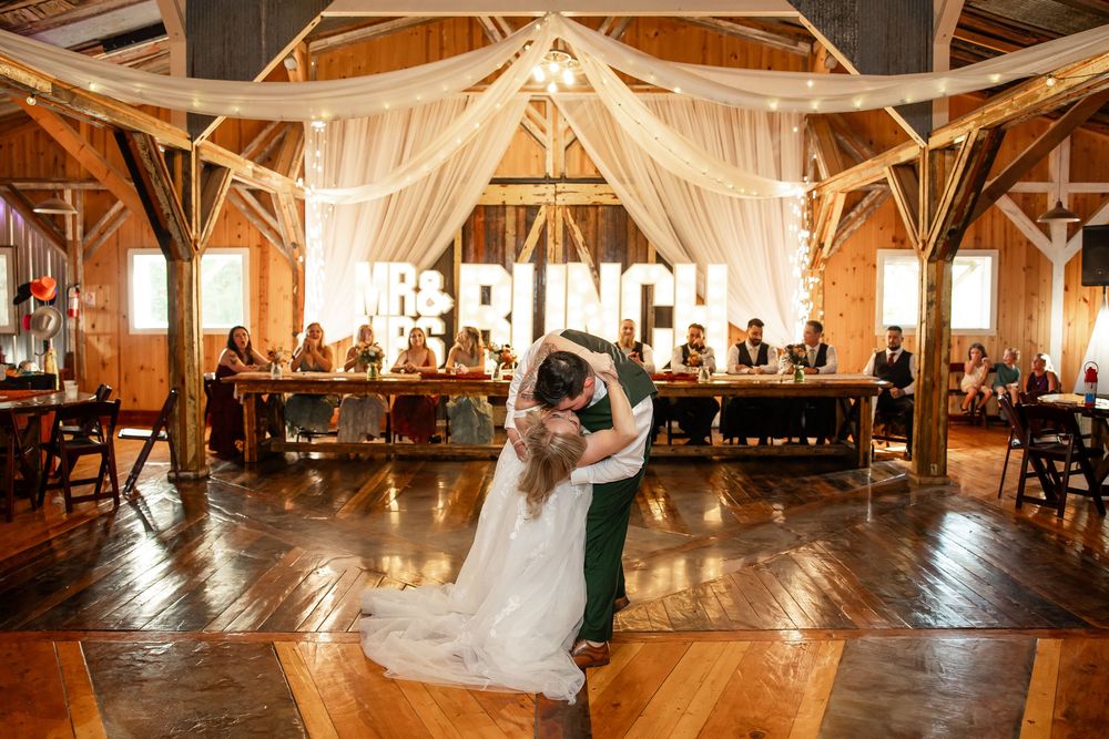Bride and groom share first dance in rustic barn wedding venue with draped fabric ceiling and Mr & Mrs sign.