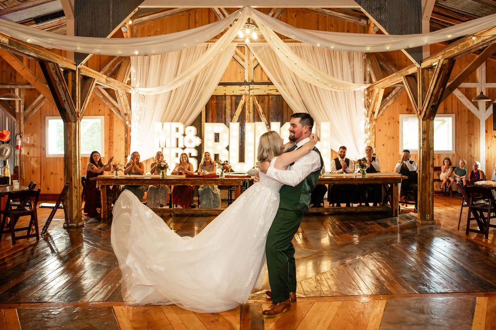 Bride and groom share first dance in rustic barn wedding venue with draped fabric decor and guests seated at tables.