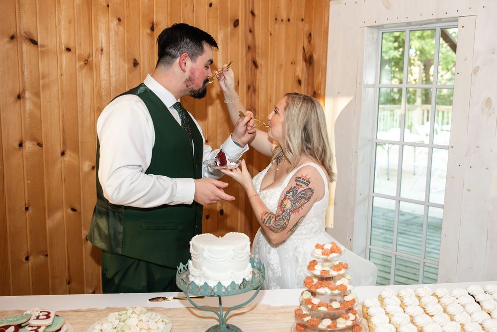 Bride feeding groom wedding cake during reception at a rustic barn venue with cupcake display.