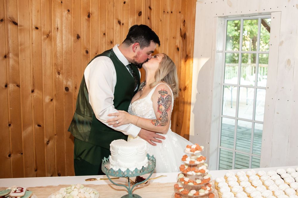 Newlyweds sharing a romantic kiss at their wedding cake table in a rustic barn venue.