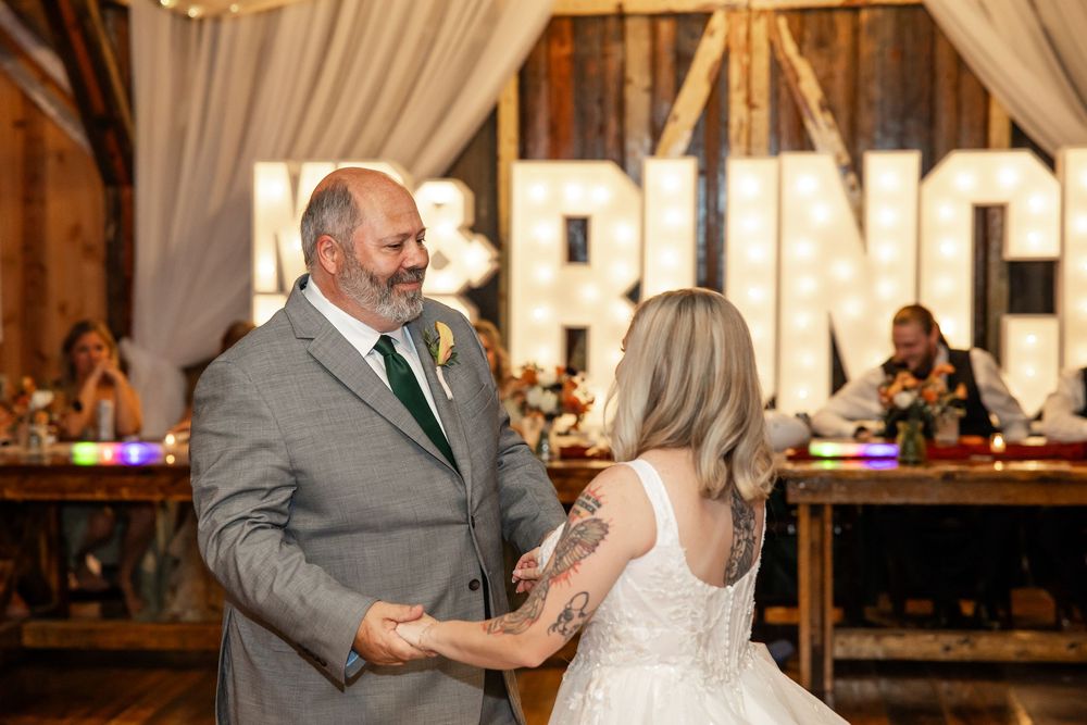 Bride and groom share first dance in rustic barn wedding venue with marquee letter backdrop.