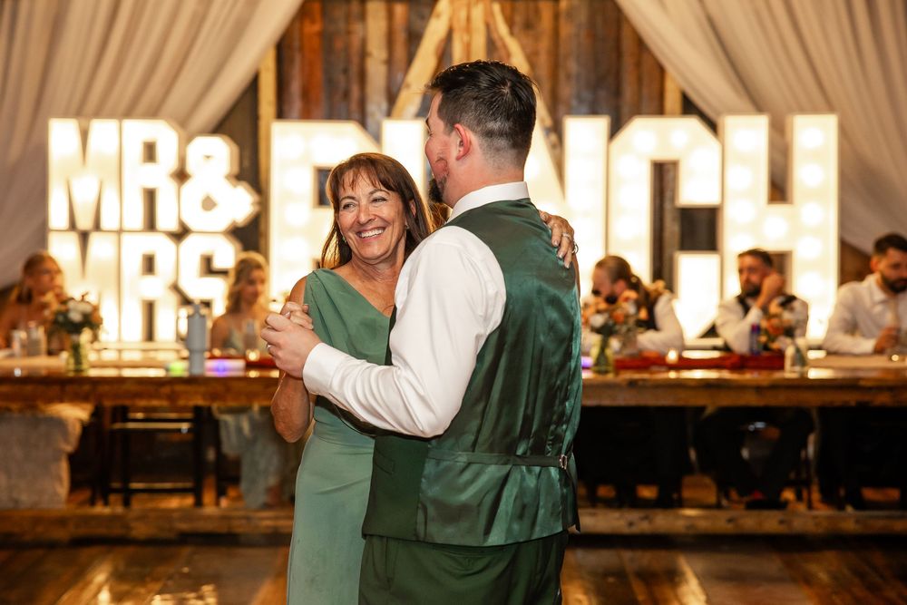 Groom and mother share a wedding dance in front of glowing MR & MRS marquee letters at elegant reception.