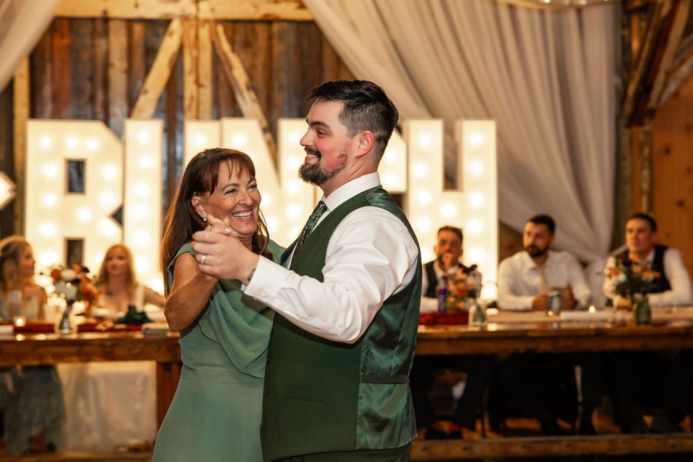 Groom in green vest shares mother-son dance at rustic barn wedding reception with marquee letters backdrop.
