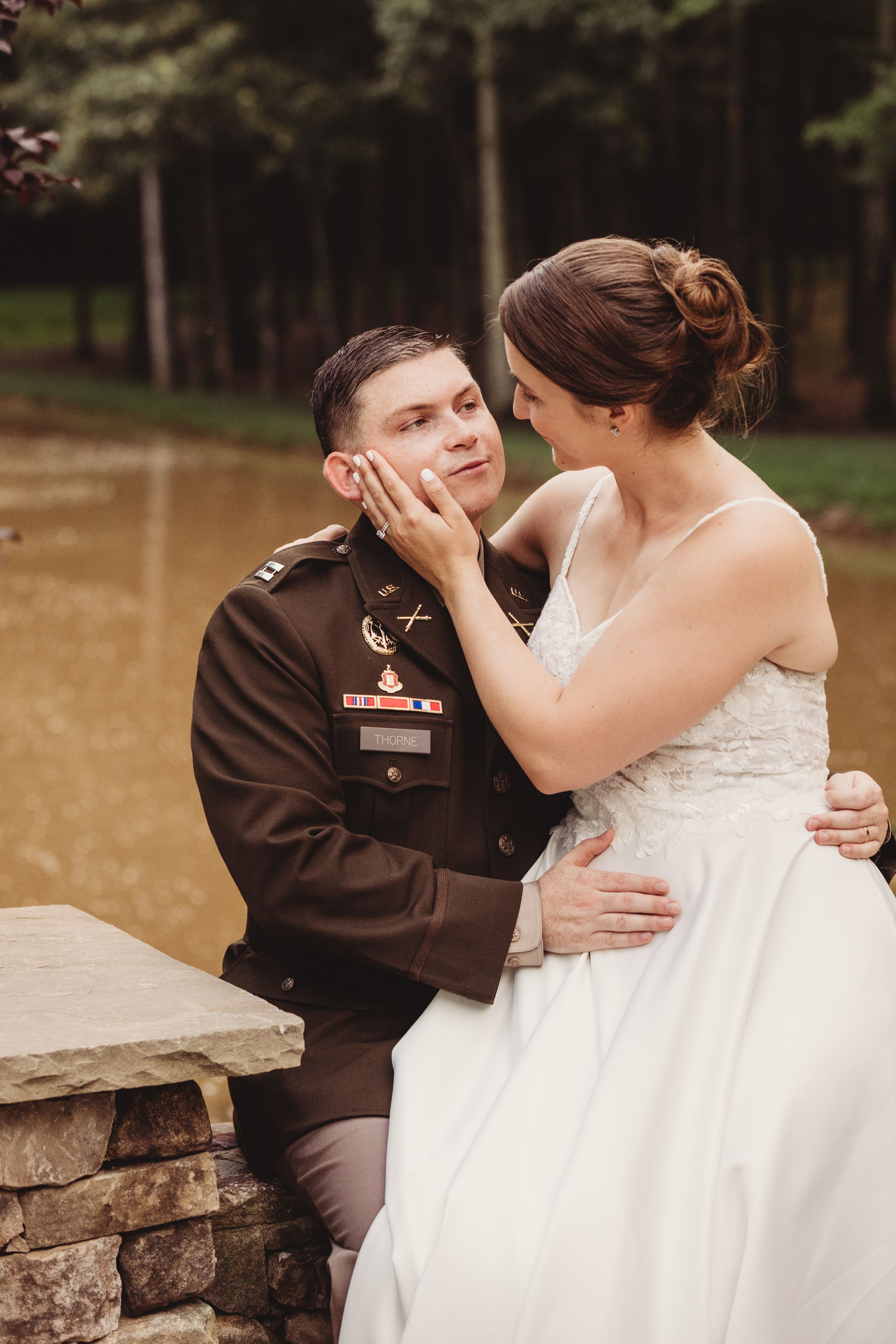 Bride in white gown tenderly cups grooms face in military uniform during romantic outdoor wedding portrait.