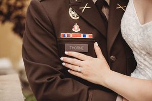 Close-up of military uniform with Thorne name tag and medals, embraced by bride wearing engagement ring.