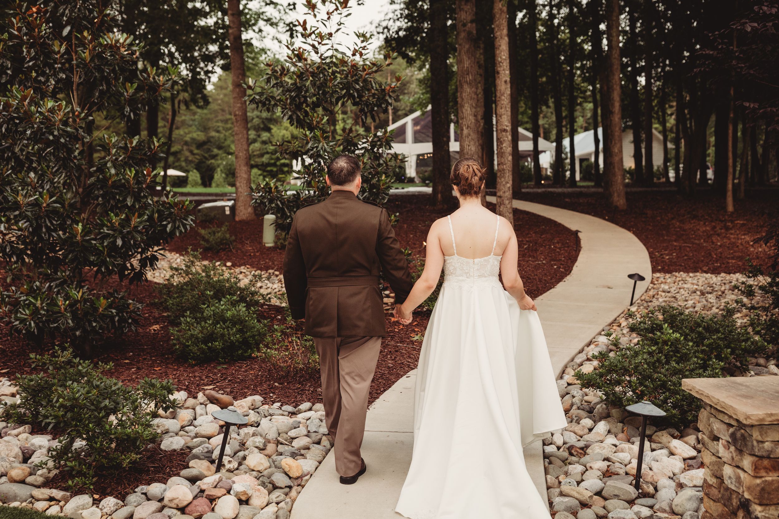Bride and groom walking hand in hand along a curved garden path surrounded by trees and decorative stones.