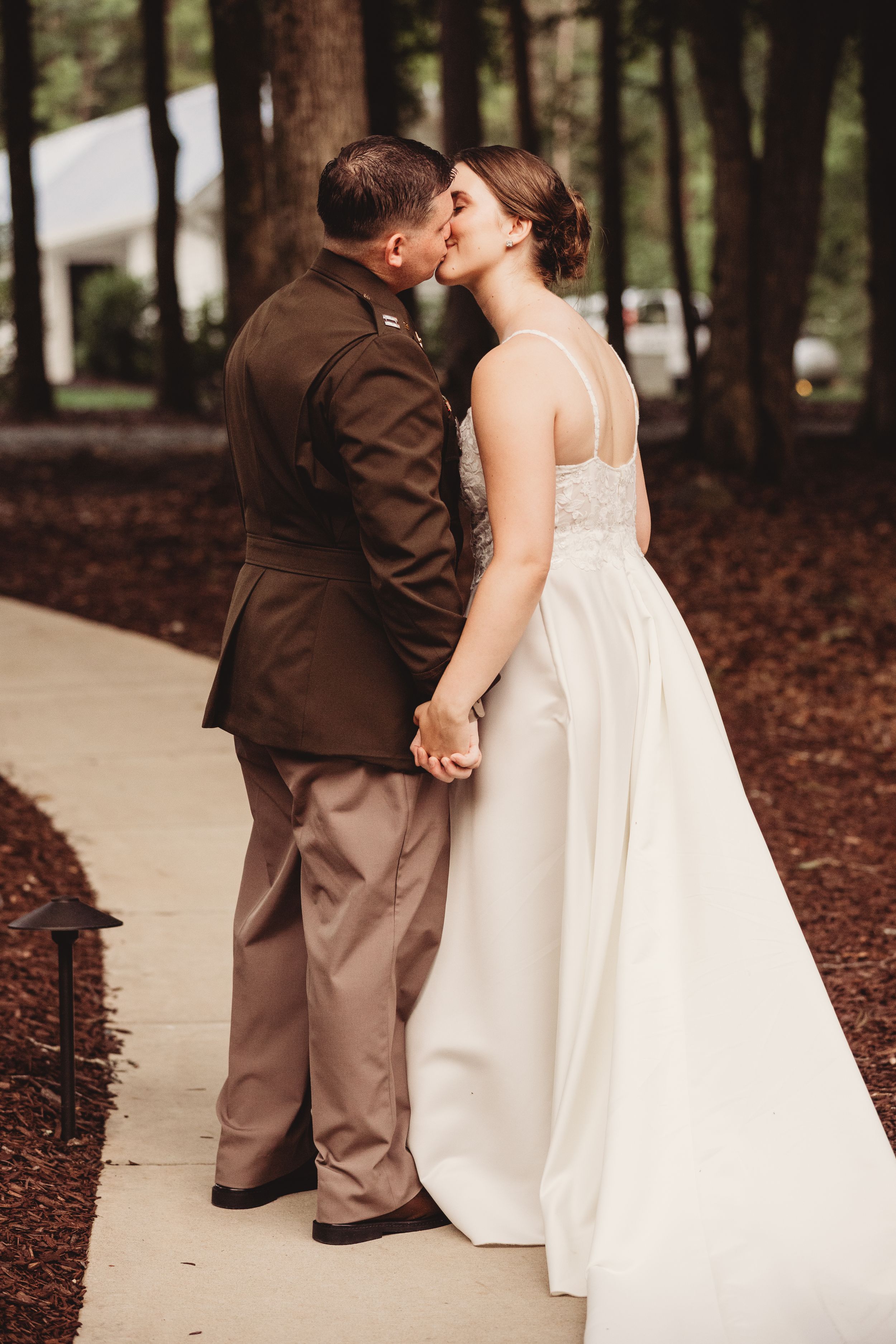 Military groom in uniform kissing bride in white wedding gown while holding hands on outdoor pathway.