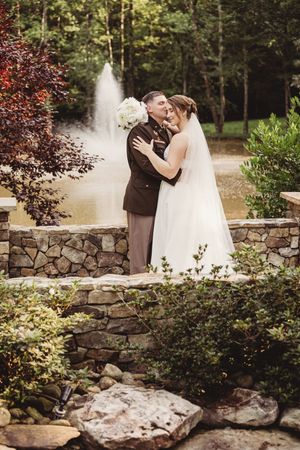 Bride and groom embrace near a stone wall with a fountain and lush greenery in a romantic outdoor wedding setting.