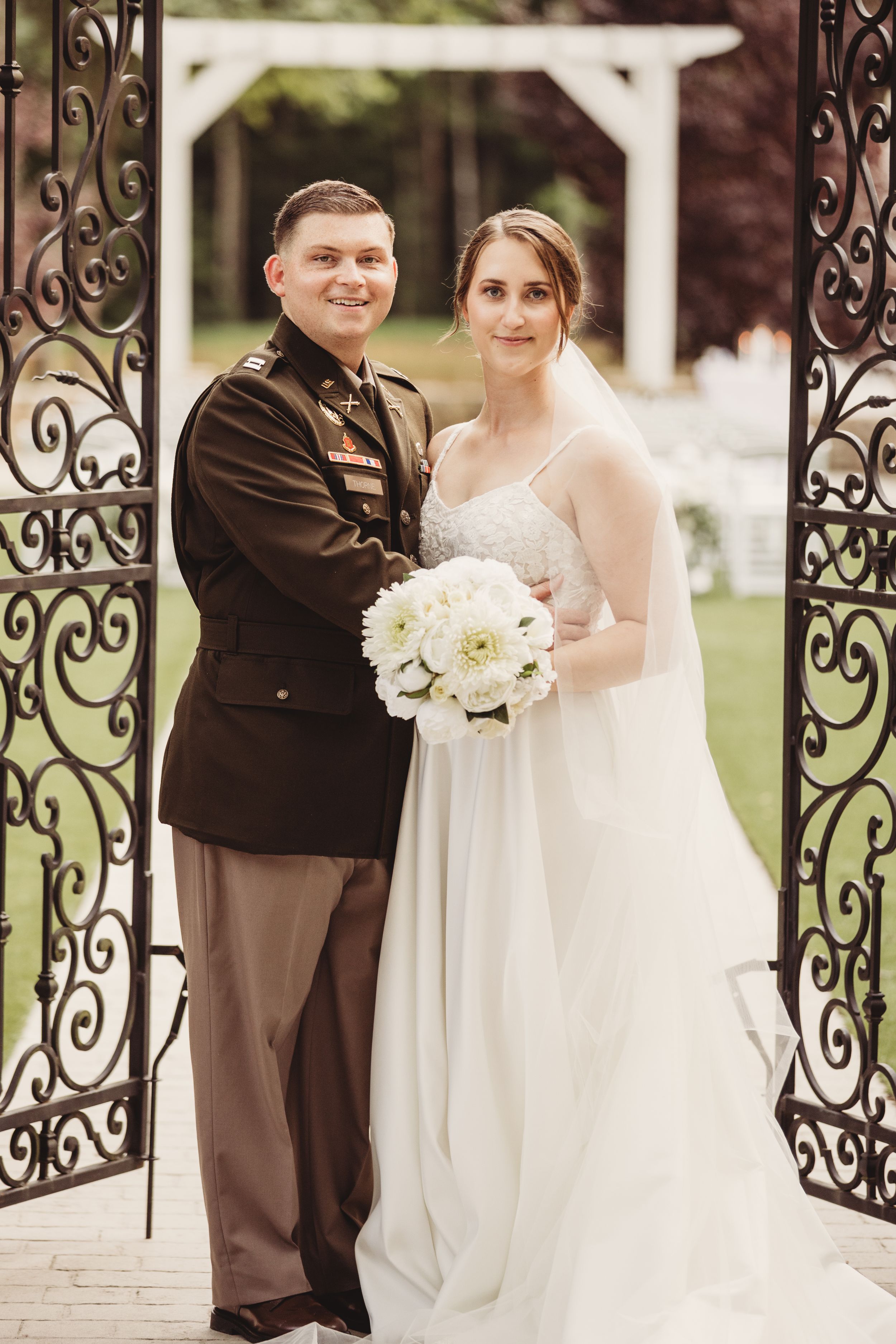 Military groom in dress uniform smiles with bride in white gown holding bouquet by ornate iron gates.