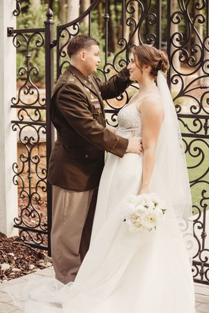Military groom in uniform tenderly touches bride's face at ornate iron gate, bride holds white bouquet.