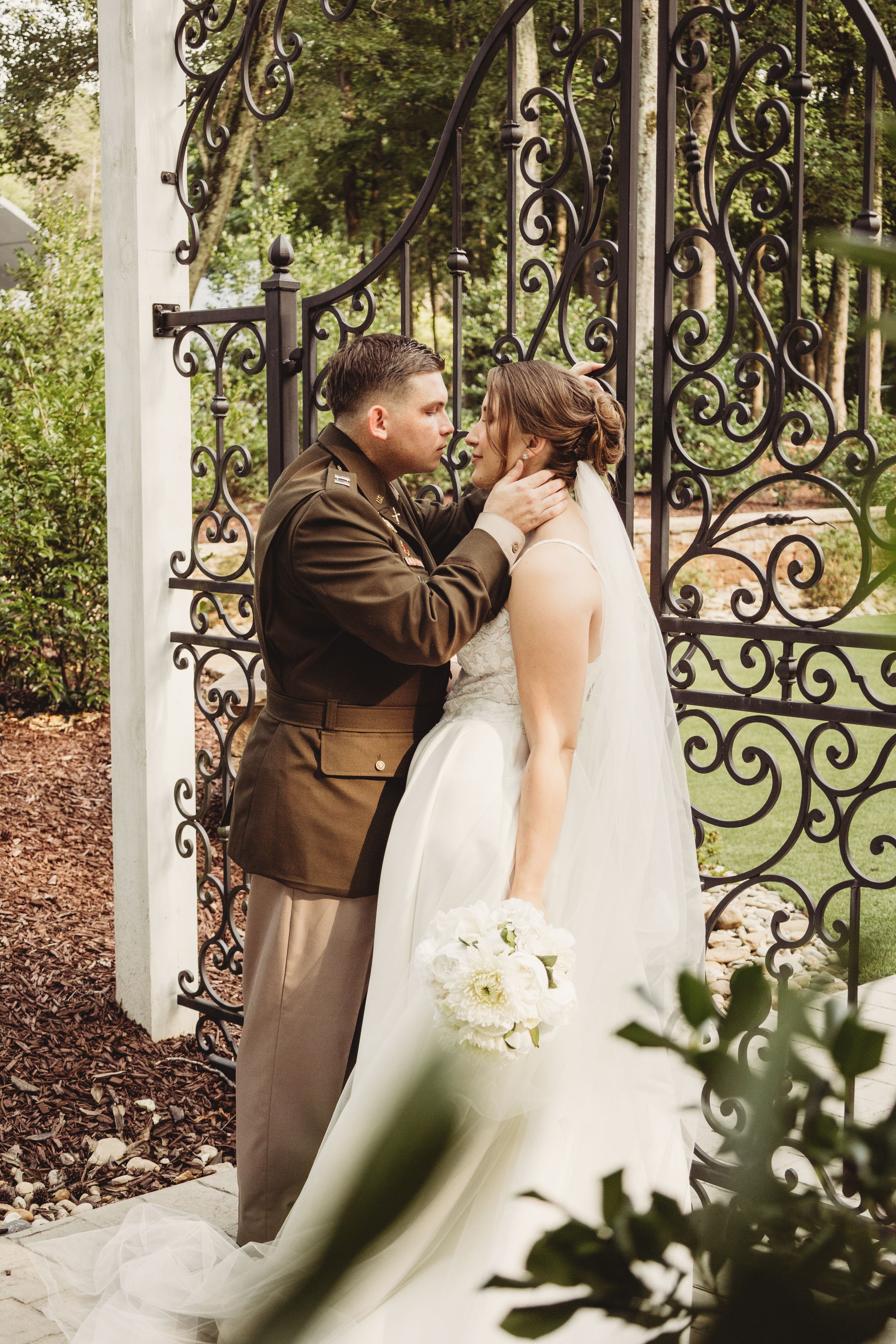 Bride and groom share a romantic moment by ornate iron garden gates, bride holding white bouquet in elegant wedding attire.