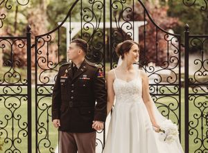 Military groom in dress uniform holds hands with bride in white gown before ornate iron garden gates.