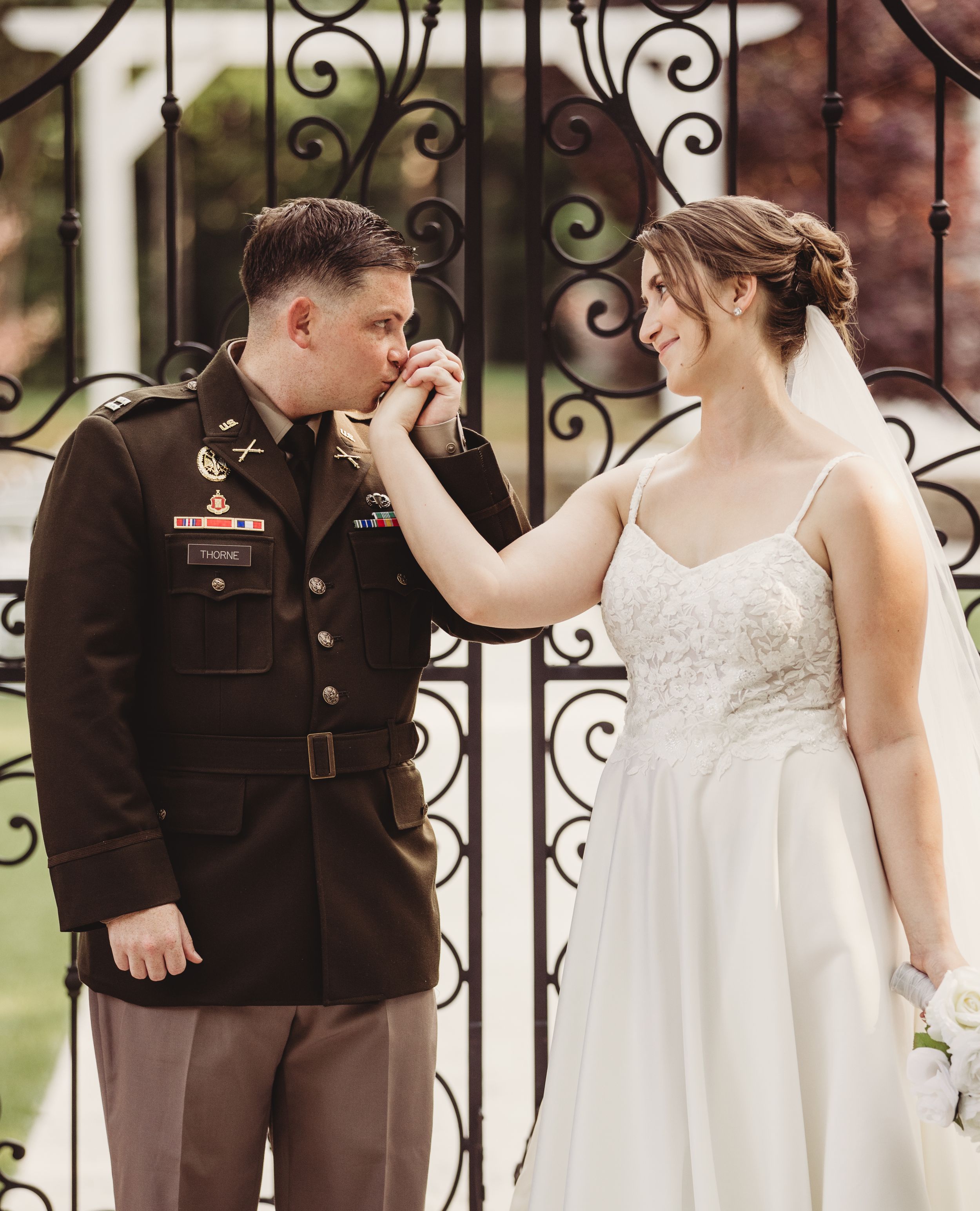 Military groom in dress uniform kisses bride's hand at ornate iron gate during romantic wedding ceremony.
