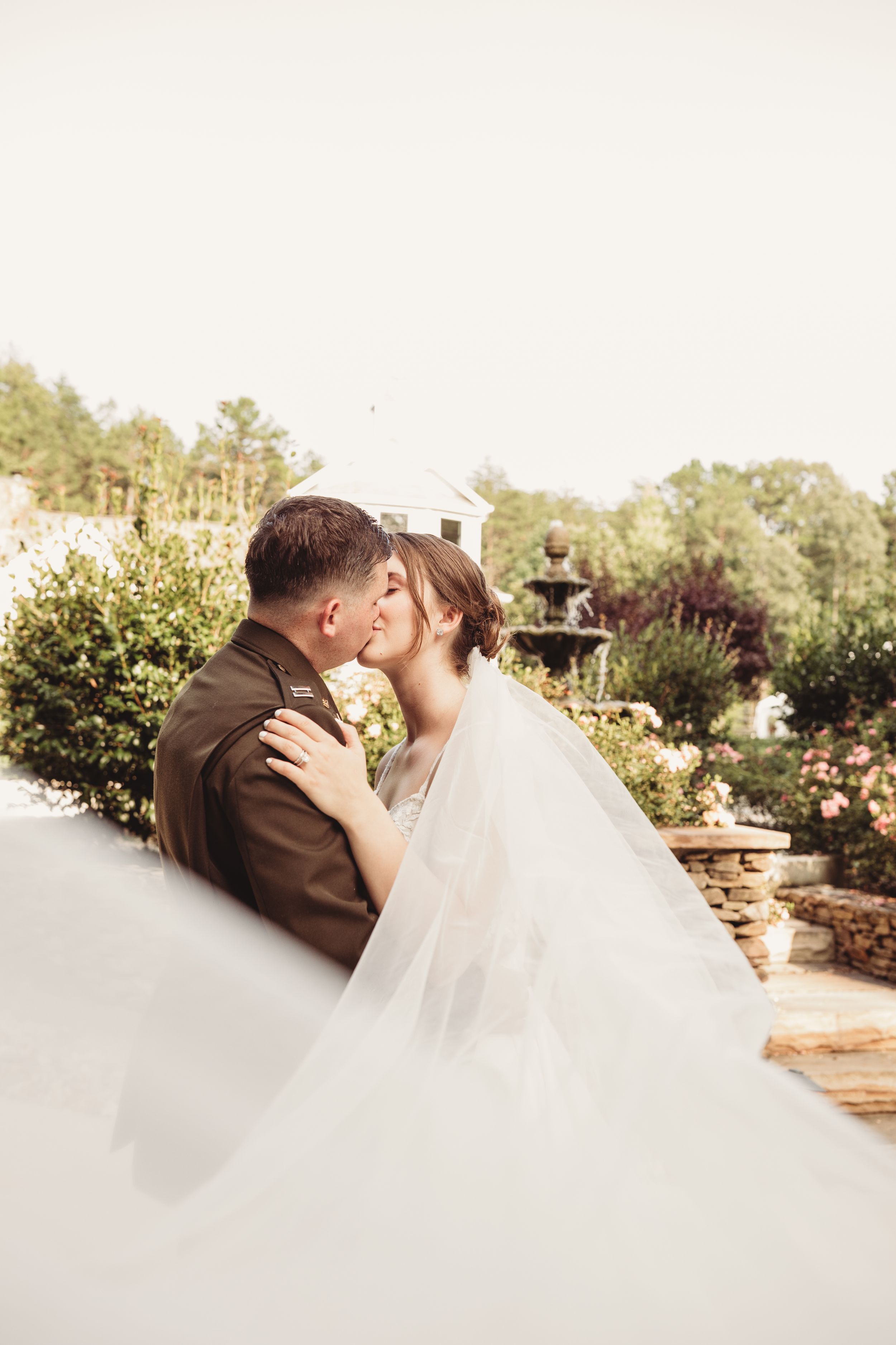 Military groom and bride share romantic kiss outdoors, veil flowing, garden fountain in background.
