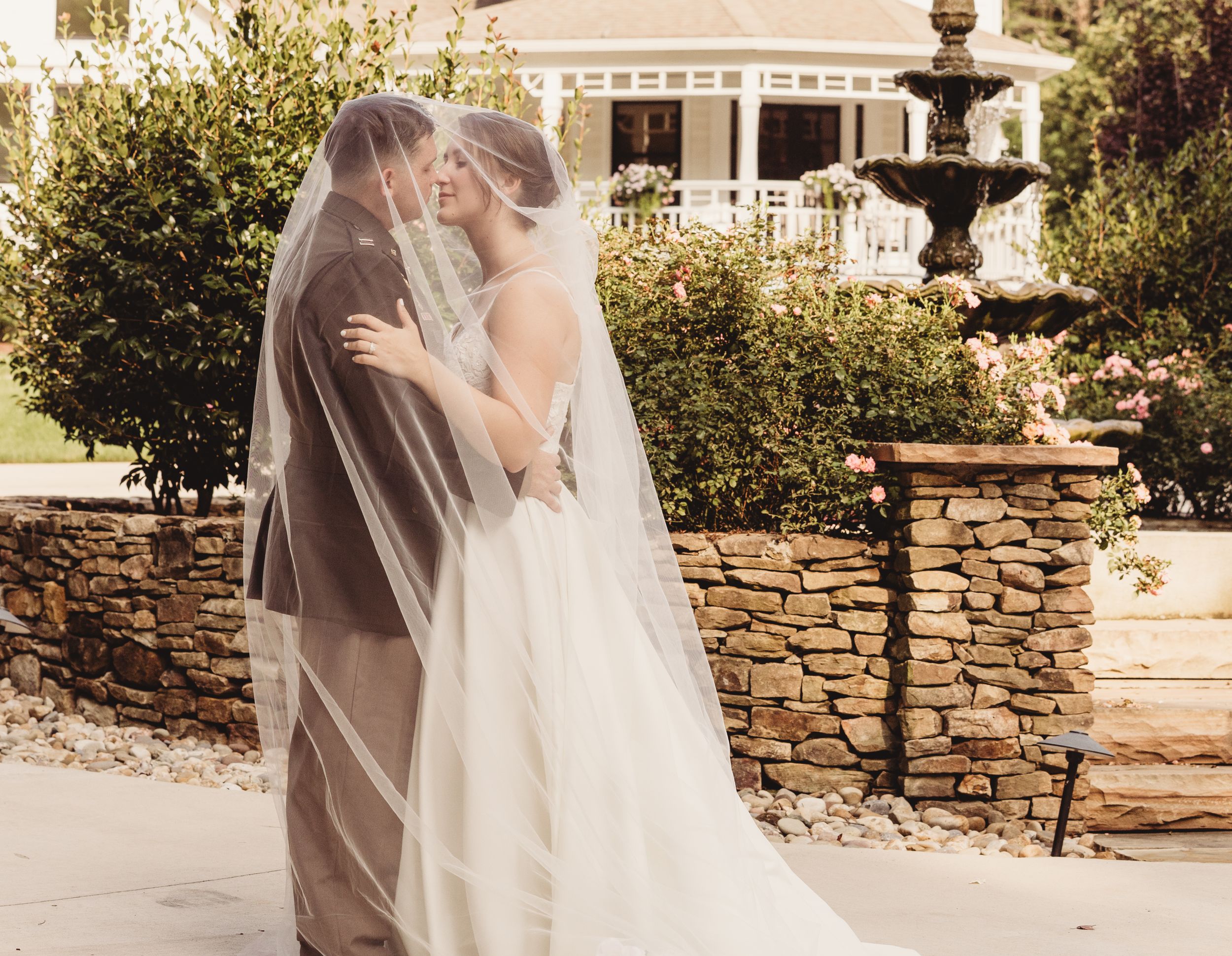 Bride and groom sharing a kiss under a flowing veil at an elegant outdoor wedding venue with a stone fountain.