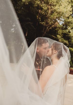 A bride and groom share a romantic kiss beneath a flowing wedding veil outdoors, with lush green trees in the background.