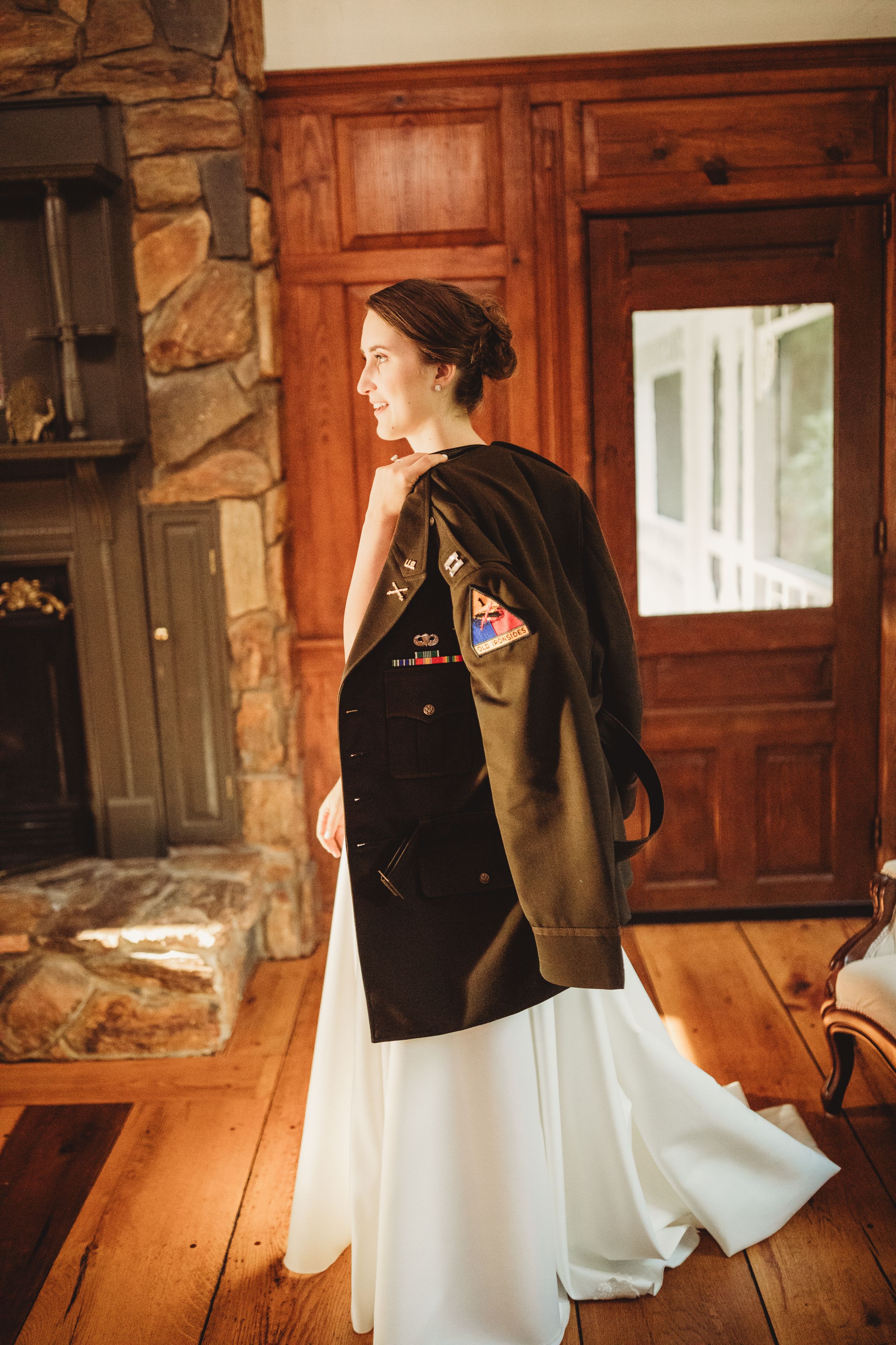 Smiling bride in white gown wearing military jacket with patches, standing in rustic wooden interior near stone fireplace.
