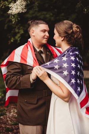 Military groom and bride wrapped together in American flag, sharing romantic moment in lush green outdoor setting.