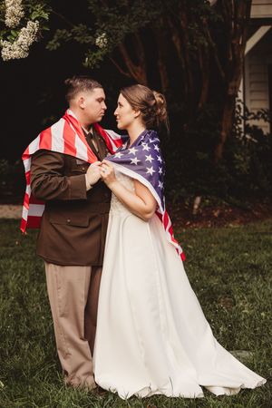Bride and groom wrapped together in an American flag, gazing at each other outdoors near blooming trees.
