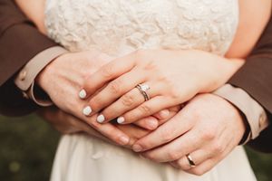 Newlywed couple holding hands showing wedding rings and engagement ring, bride in white lace dress.