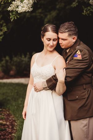 Groom in military uniform embraces bride in white lace dress outdoors among blooming trees.