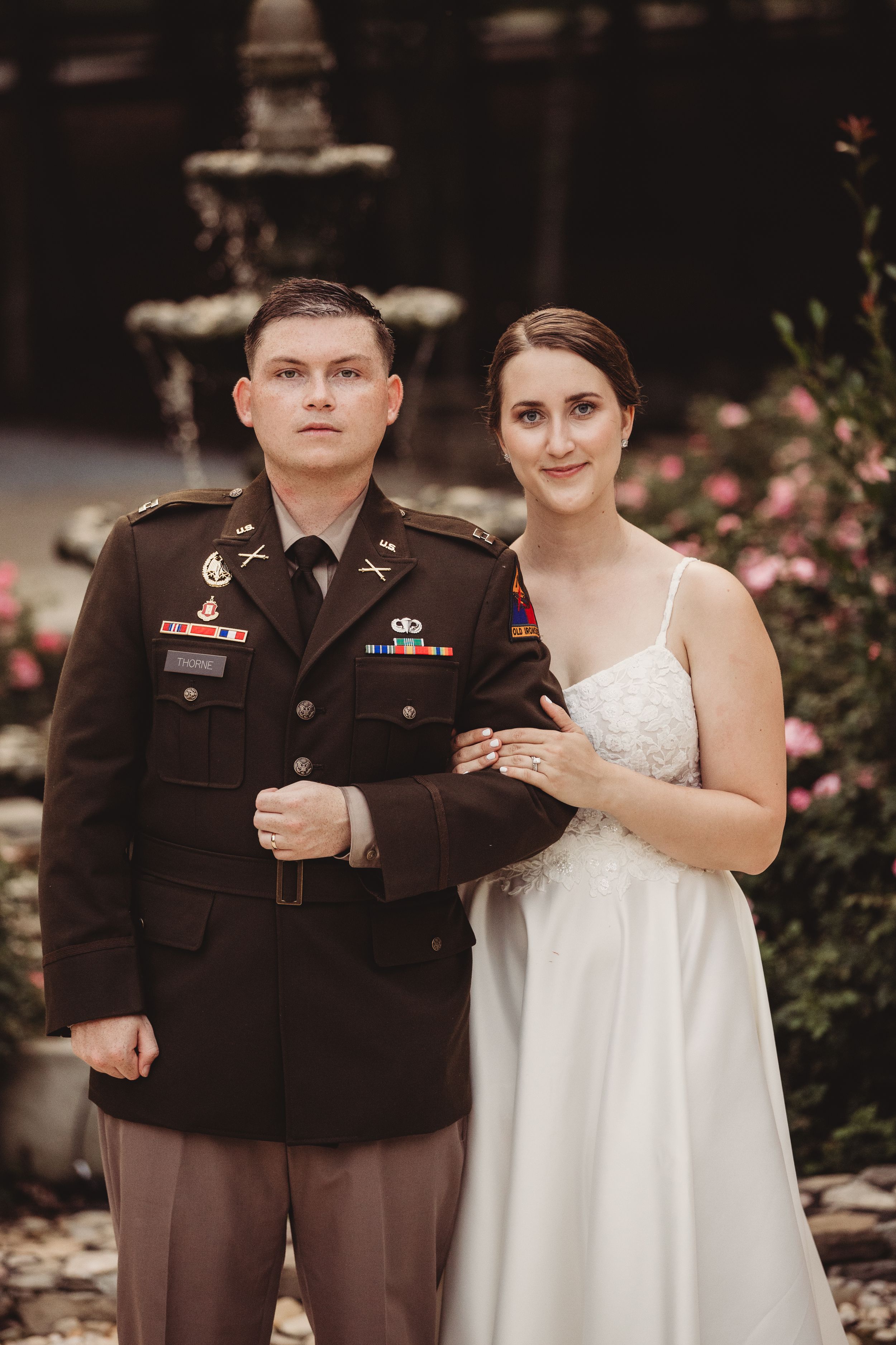 Military groom in dress uniform stands with bride in white gown outdoors near fountain and pink flowers.