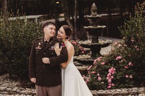 Military groom in dress uniform stands with bride in white gown by a garden fountain surrounded by pink roses.