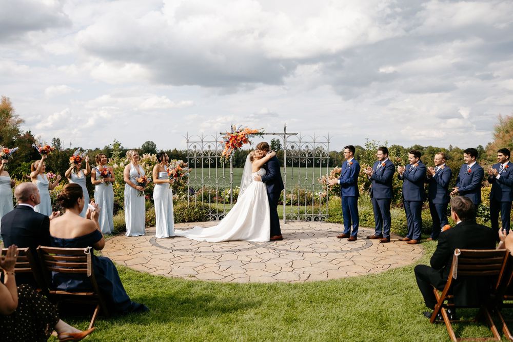 Bride and groom share first kiss at outdoor wedding ceremony with bridal party on lush green lawn.