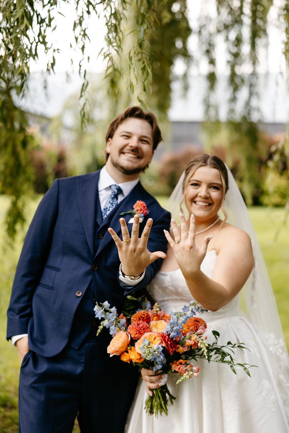 Bride and groom smiling and showing wedding rings, holding colorful bouquet under willow tree.