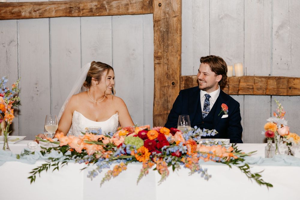 Bride and groom smile at sweetheart table adorned with vibrant orange, pink, and blue floral centerpieces at wedding reception.
