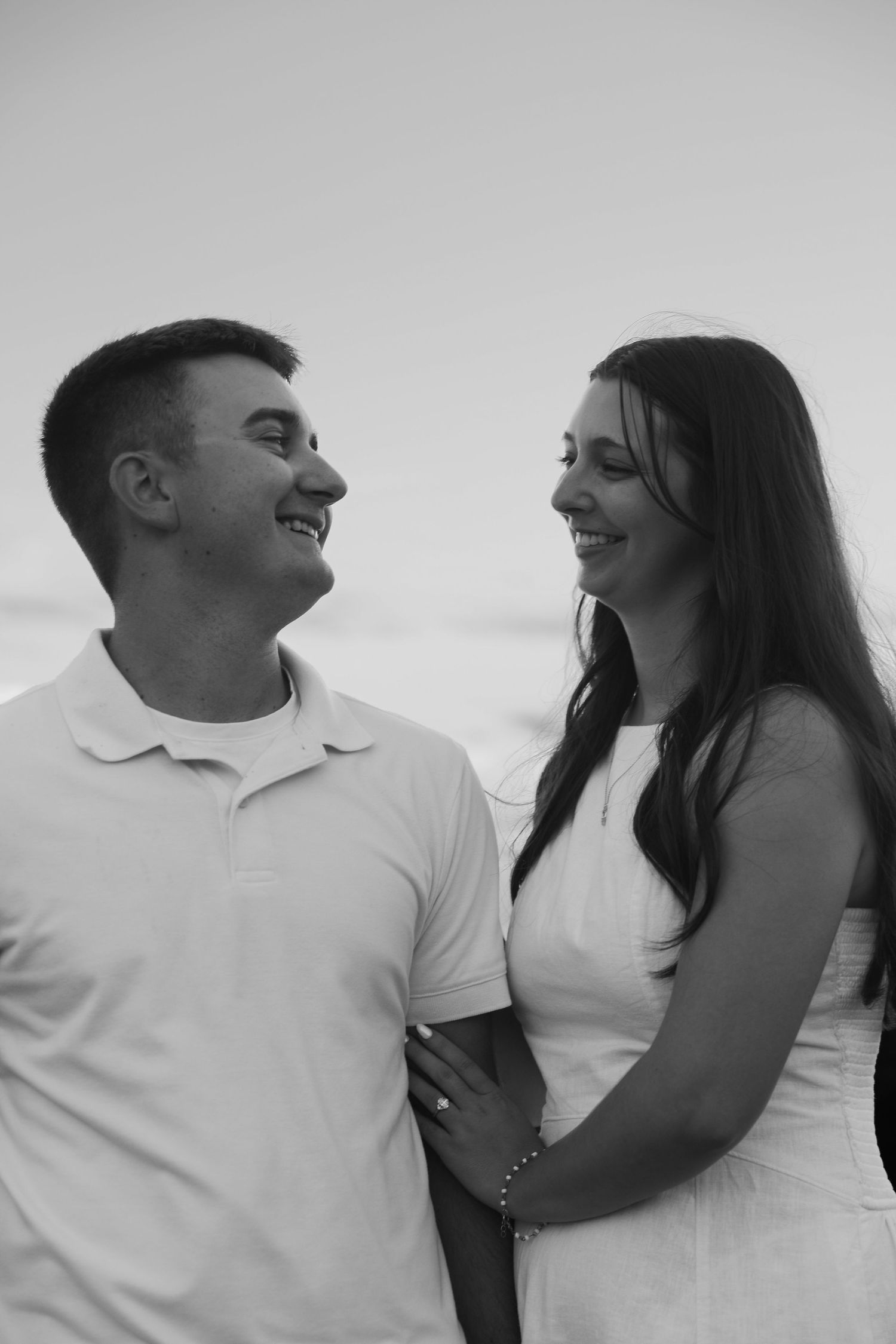 Black and white photo of a smiling couple gazing at each other outdoors, woman in white dress, man in polo shirt.