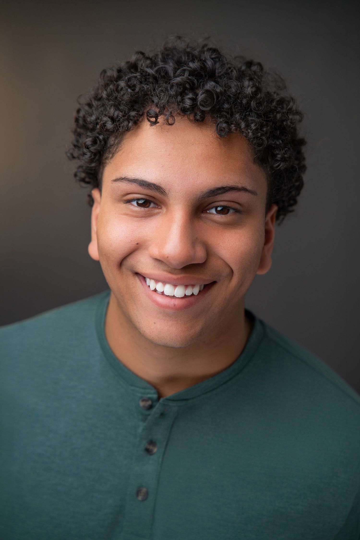 Young man with curly hair smiling confidently in a teal henley shirt against a gray background.
