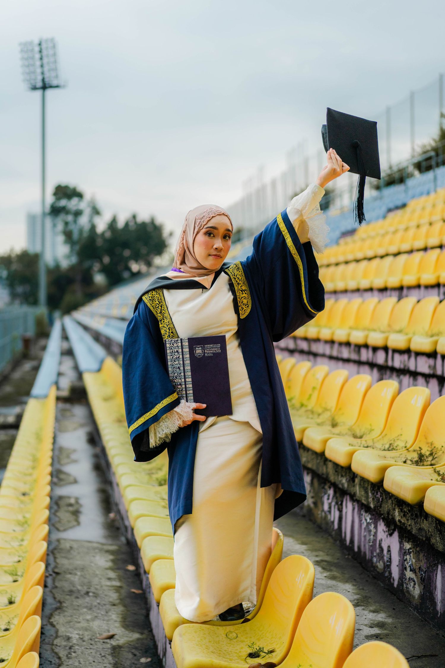 Muslim woman in graduation gown and hijab holds diploma, raises cap among yellow stadium seats.