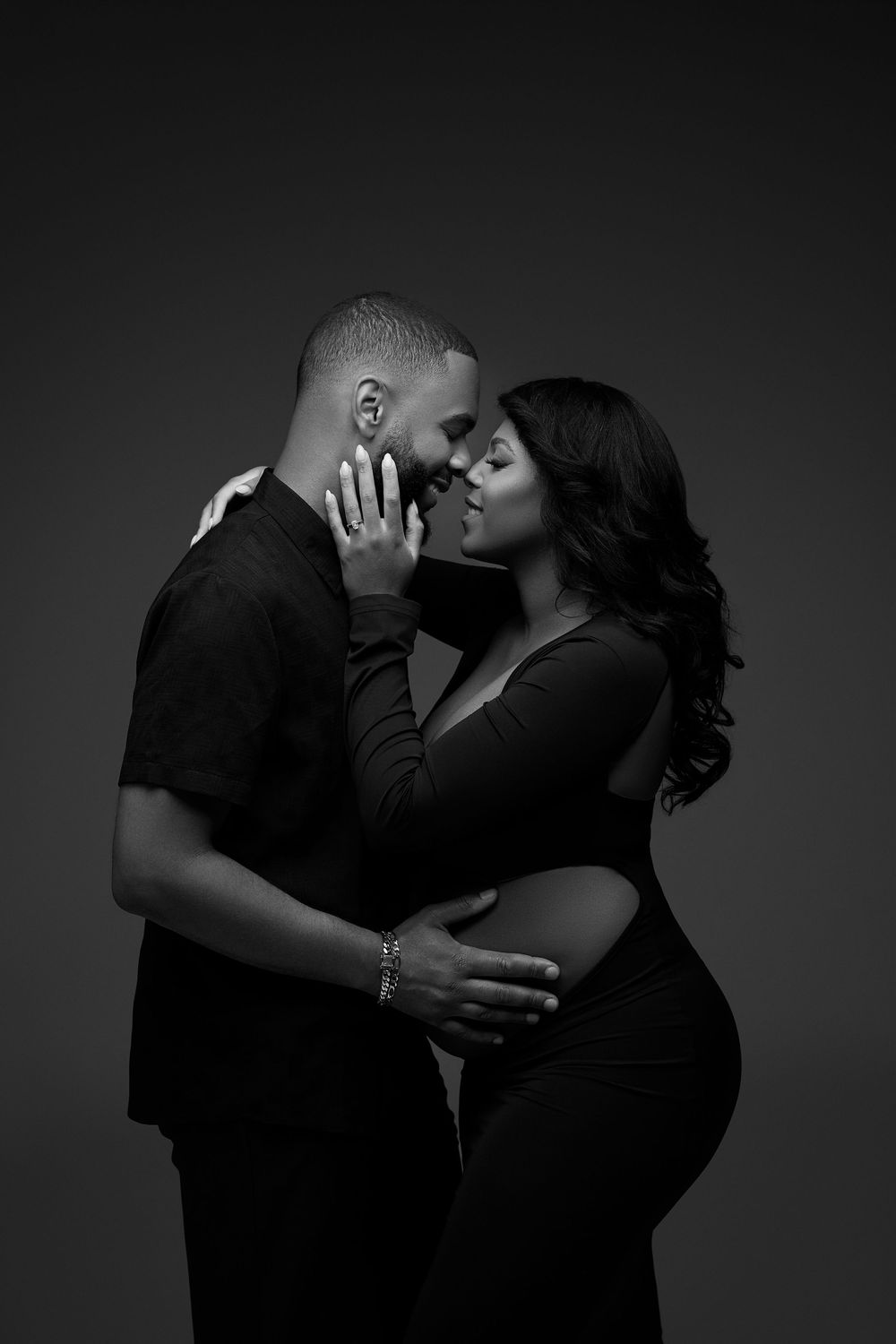 Black and white maternity photo of a couple embracing, man cradling pregnant womans belly while almost kissing.
