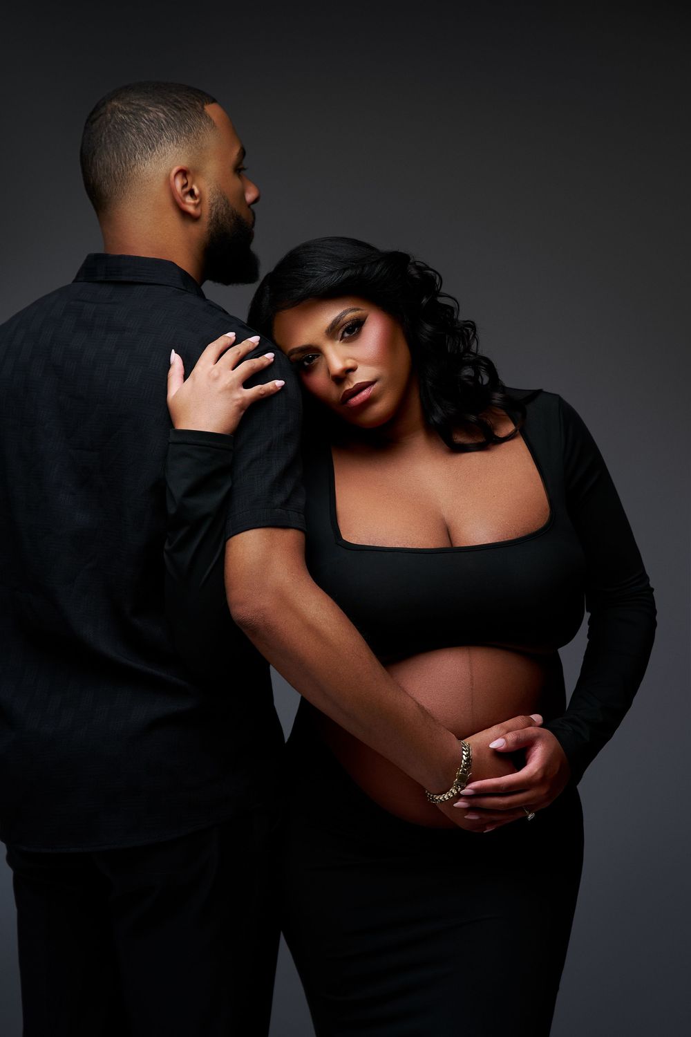 A pregnant woman in black crop top leans against her partner in a studio maternity photo shoot.