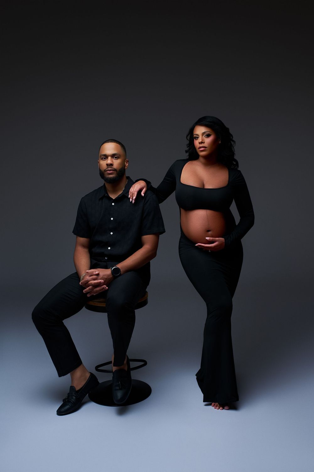 A pregnant couple in matching black outfits poses in a studio, the man seated and woman standing, cradling her baby bump.