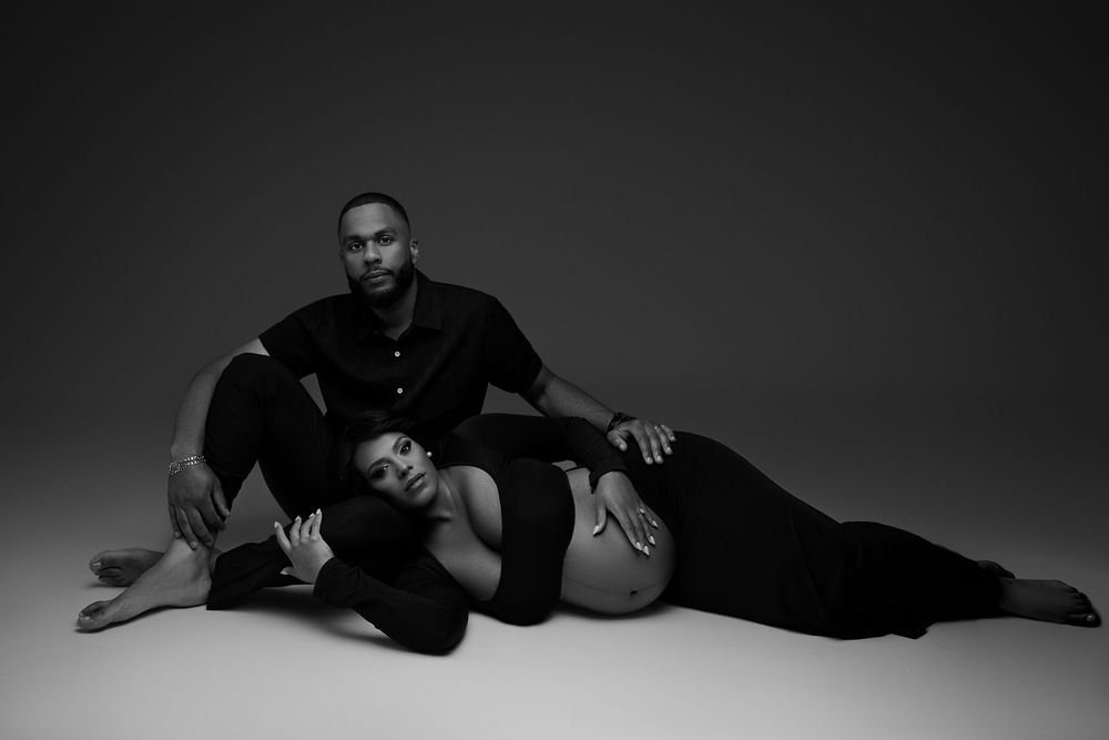 Black and white maternity photo of a pregnant woman lying down with a man seated behind her on a studio floor.