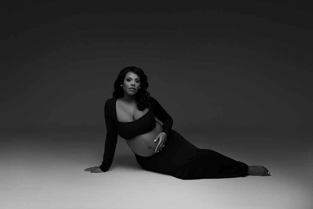 Black and white maternity photo of a pregnant woman in a black crop top and skirt posing gracefully on a studio floor.