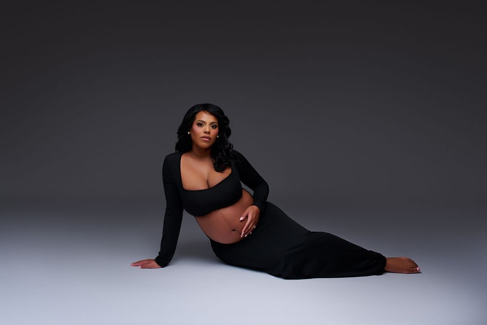 Pregnant woman in a black crop top and skirt posing elegantly on a gray studio floor in a maternity photoshoot.
