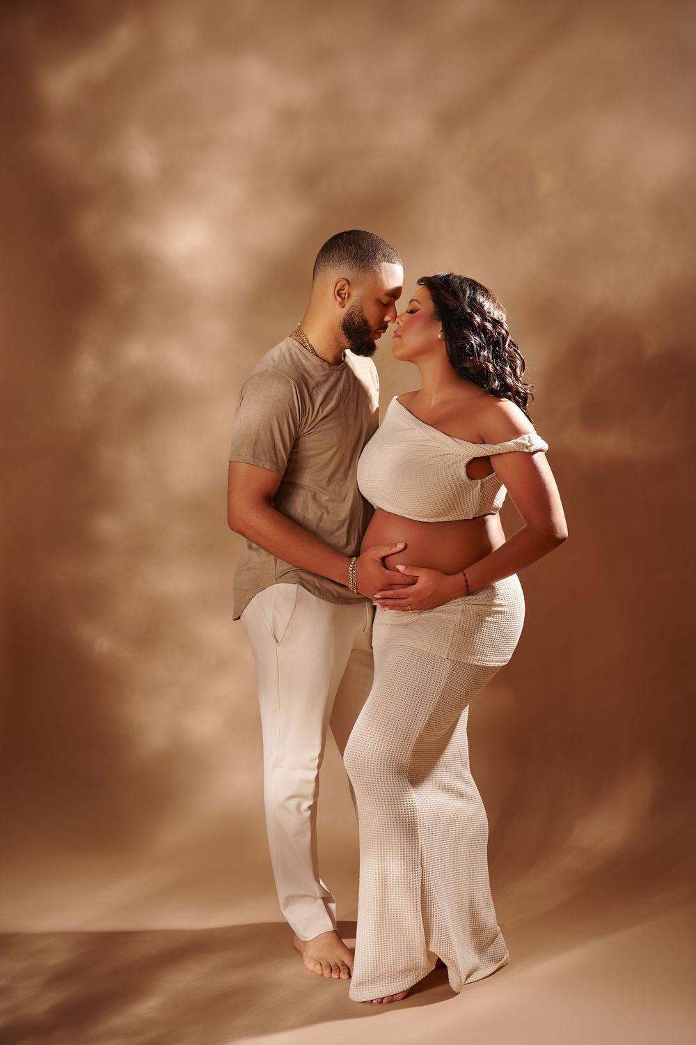 Couple in white outfits embracing in studio, pregnant woman cradling baby bump with partner kissing her cheek.