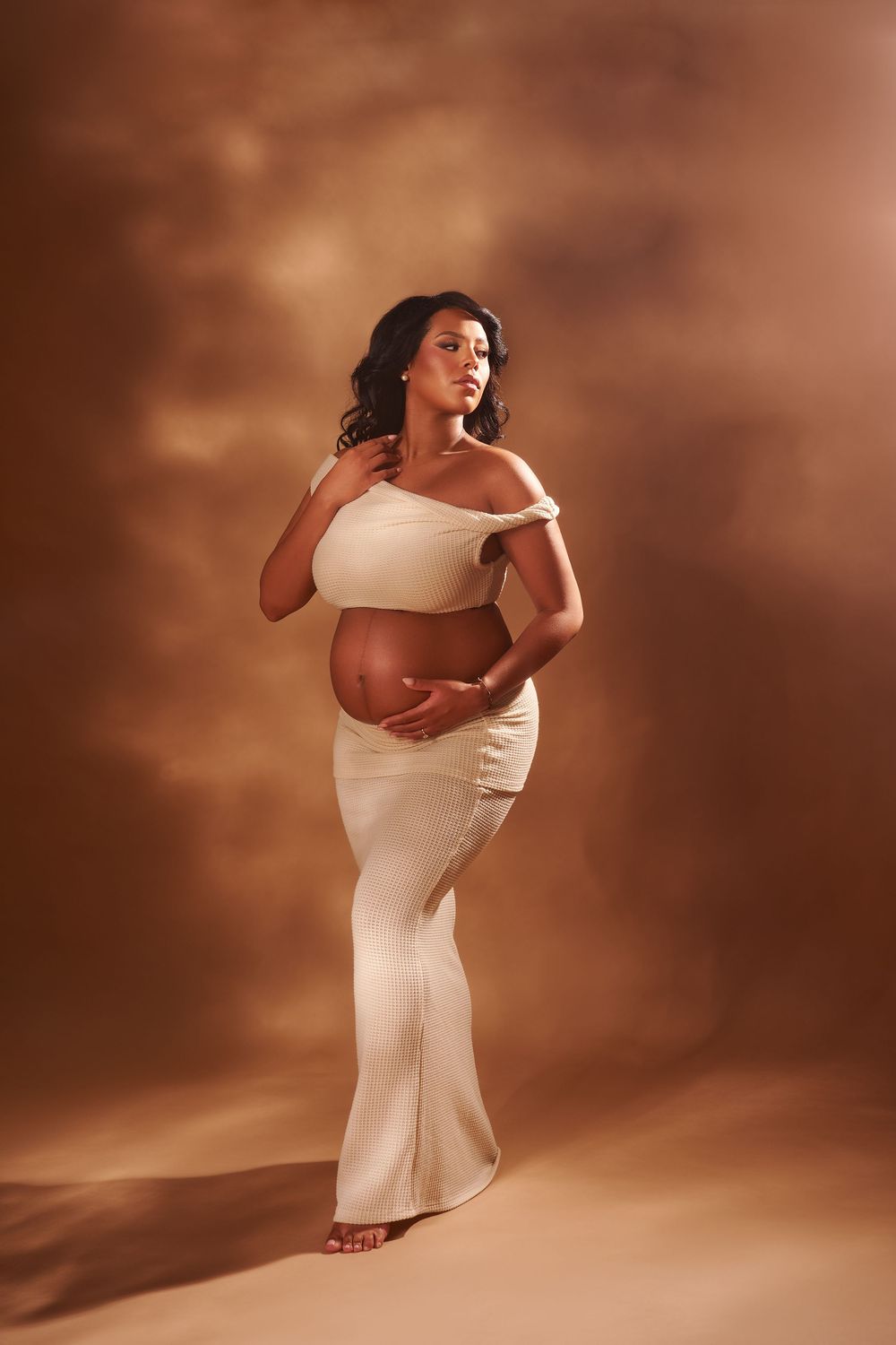 A pregnant woman poses elegantly in a white off-shoulder gown against a warm brown studio backdrop.