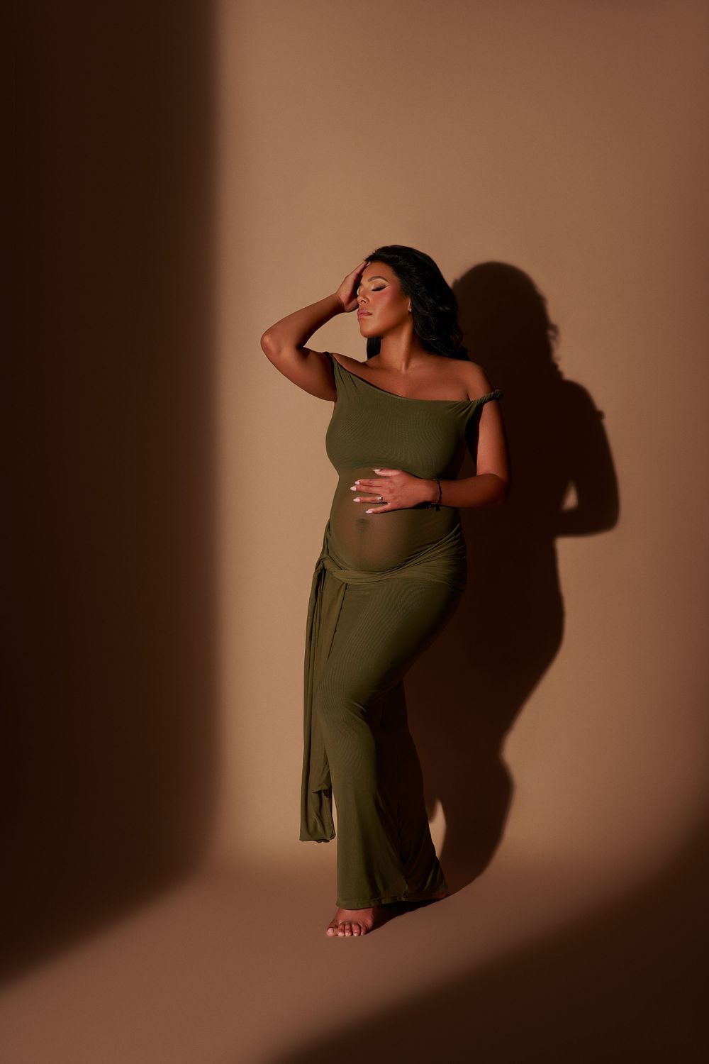 Pregnant woman in olive green one-shoulder jumpsuit poses gracefully against warm beige backdrop with dramatic shadows.