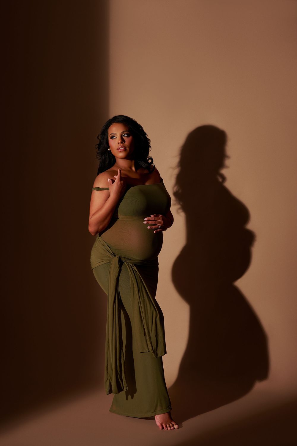 Pregnant woman in olive green wrap dress poses gracefully in studio with dramatic shadow on warm brown background.