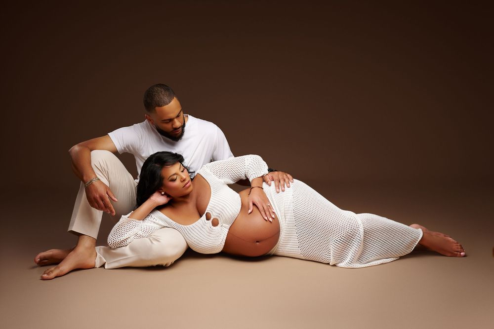 Couple in matching white outfits pose together, woman lying down cradling pregnant belly during maternity shoot.