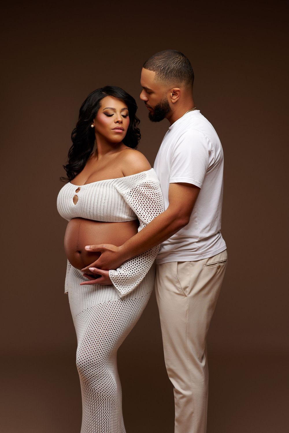 Couple in white attire posing in studio, man tenderly holding pregnant womans baby bump from behind.