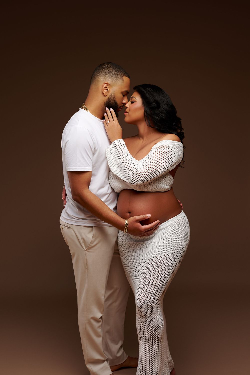 A pregnant couple in white outfits share an intimate moment, with the woman cradling her baby bump against a brown background.