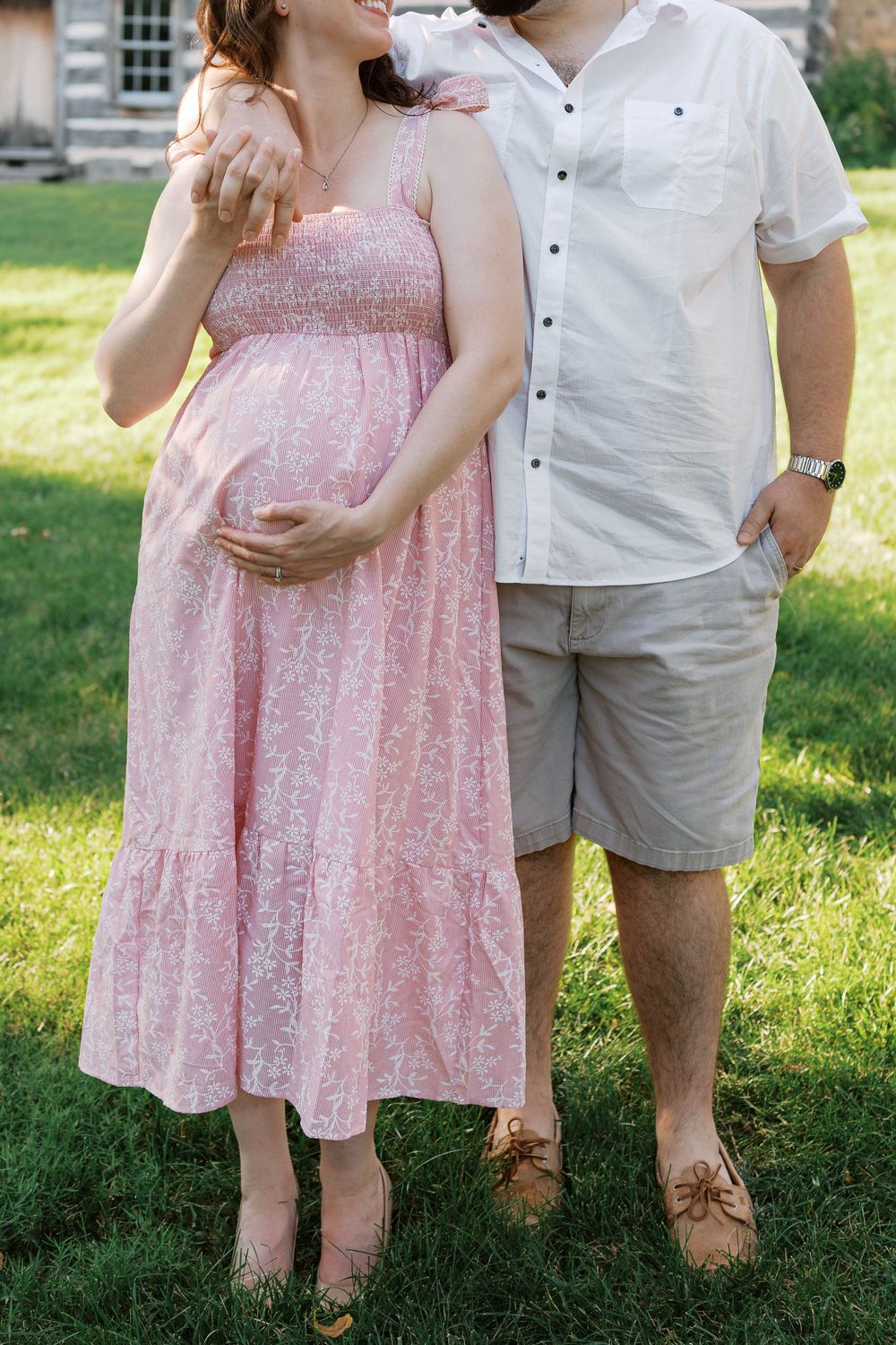 Pregnant woman in pink floral dress cradling baby bump while standing with partner on green lawn.