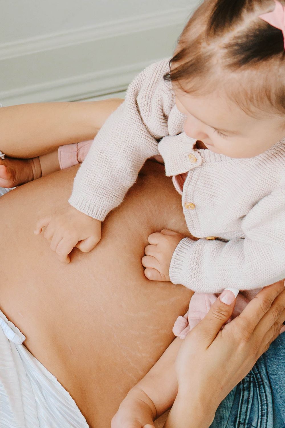 Baby girl in pink knit sweater resting on mothers pregnant belly with stretch marks.
