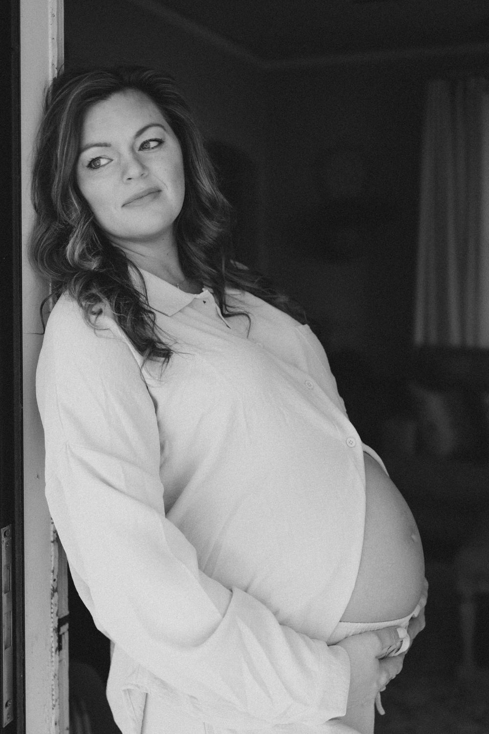 Black and white portrait of a pregnant woman cradling her belly while leaning in a doorway.